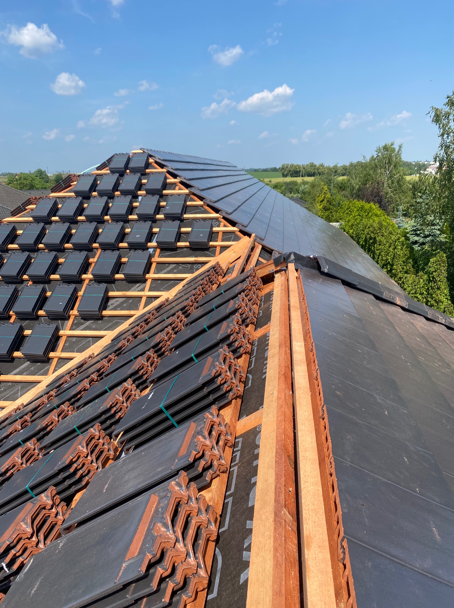 Częściowo pokryty dach z drewnianą konstrukcją, widoczne czarne dachówki ceramiczne, niektóre jeszcze w paczkach, oraz fragmenty już ułożone na dachu, w tle zieleń i błękitne niebo.