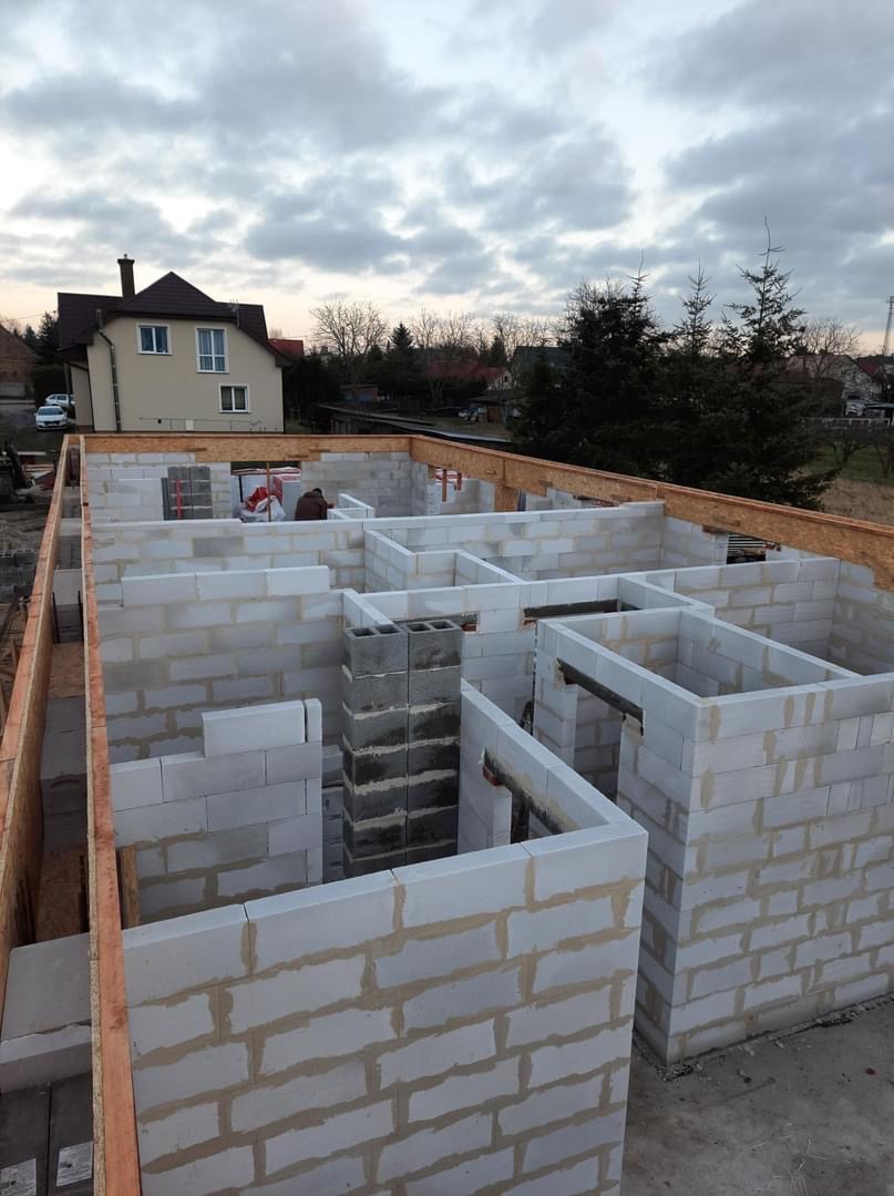 Wznoszenie ścian parteru z betonu komórkowego Ytong, widoczny komin z szarych bloczków oraz drewniane szalunki stropu, w tle dom jednorodzinny i pochmurne niebo.