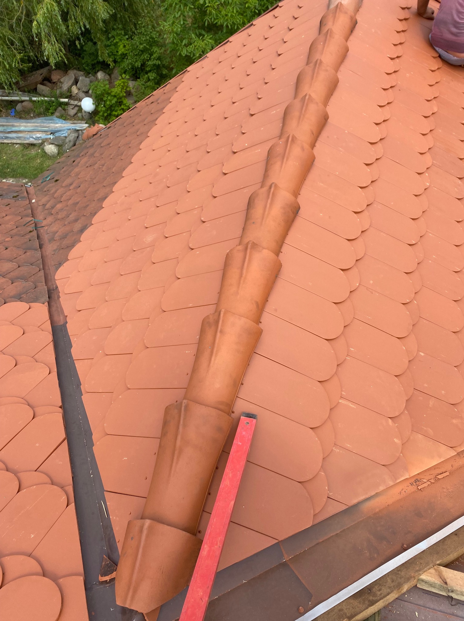 Układanie ceramicznej dachówki karpiówki w kolorze ceglastym, widoczny fragment kalenicy i poziomica, praca w toku.
