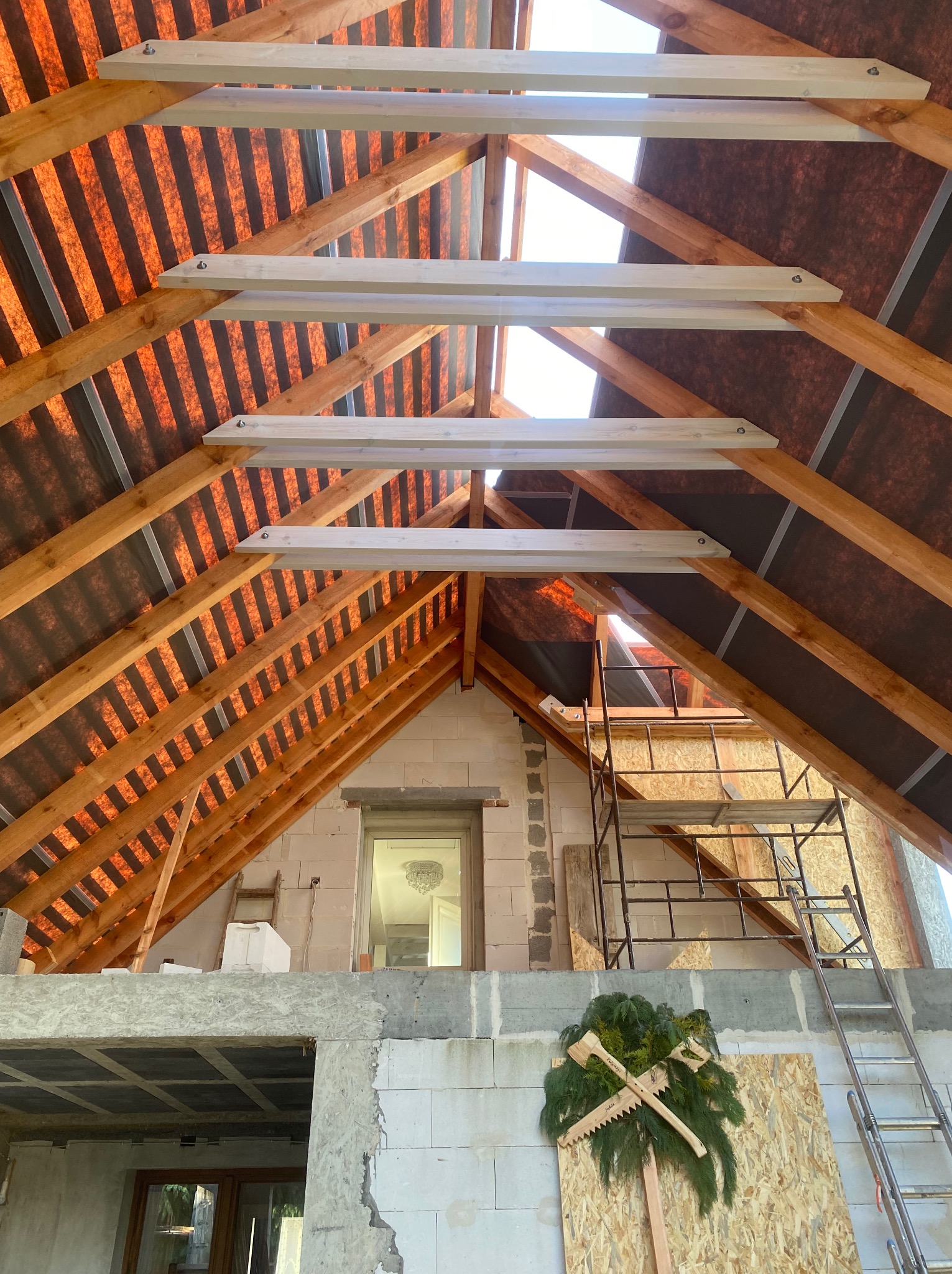 Wnętrze budowanego domu z widoczną konstrukcją dachu z drewnianymi belkami i membraną dachową, na ścianie wisi ozdoba z zielonych gałązek i drewnianych pił.