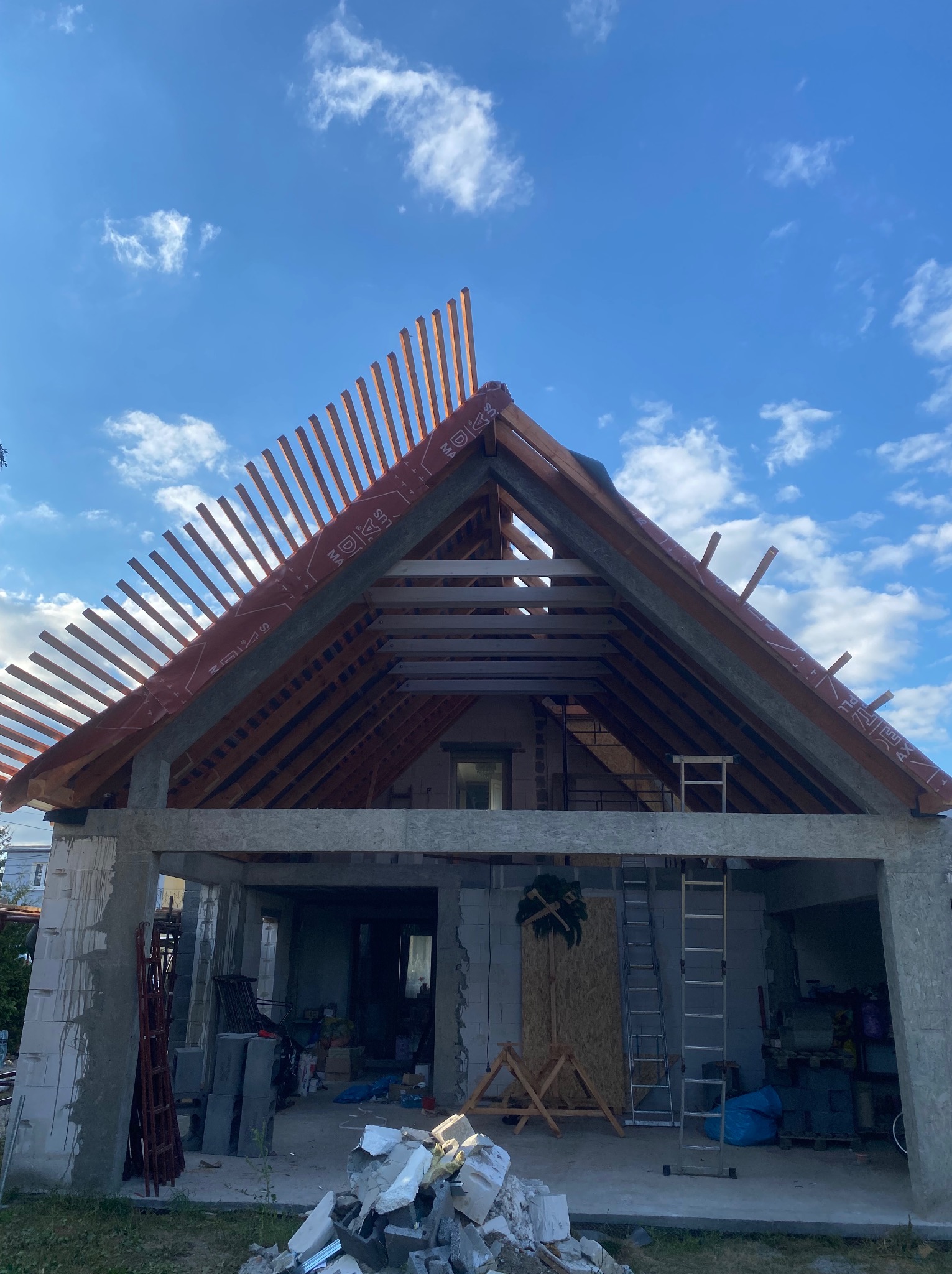 Wznoszona konstrukcja dachu domu w stanie surowym otwartym, widoczne krokwie, łaty i membrana dachowa na tle błękitnego nieba.