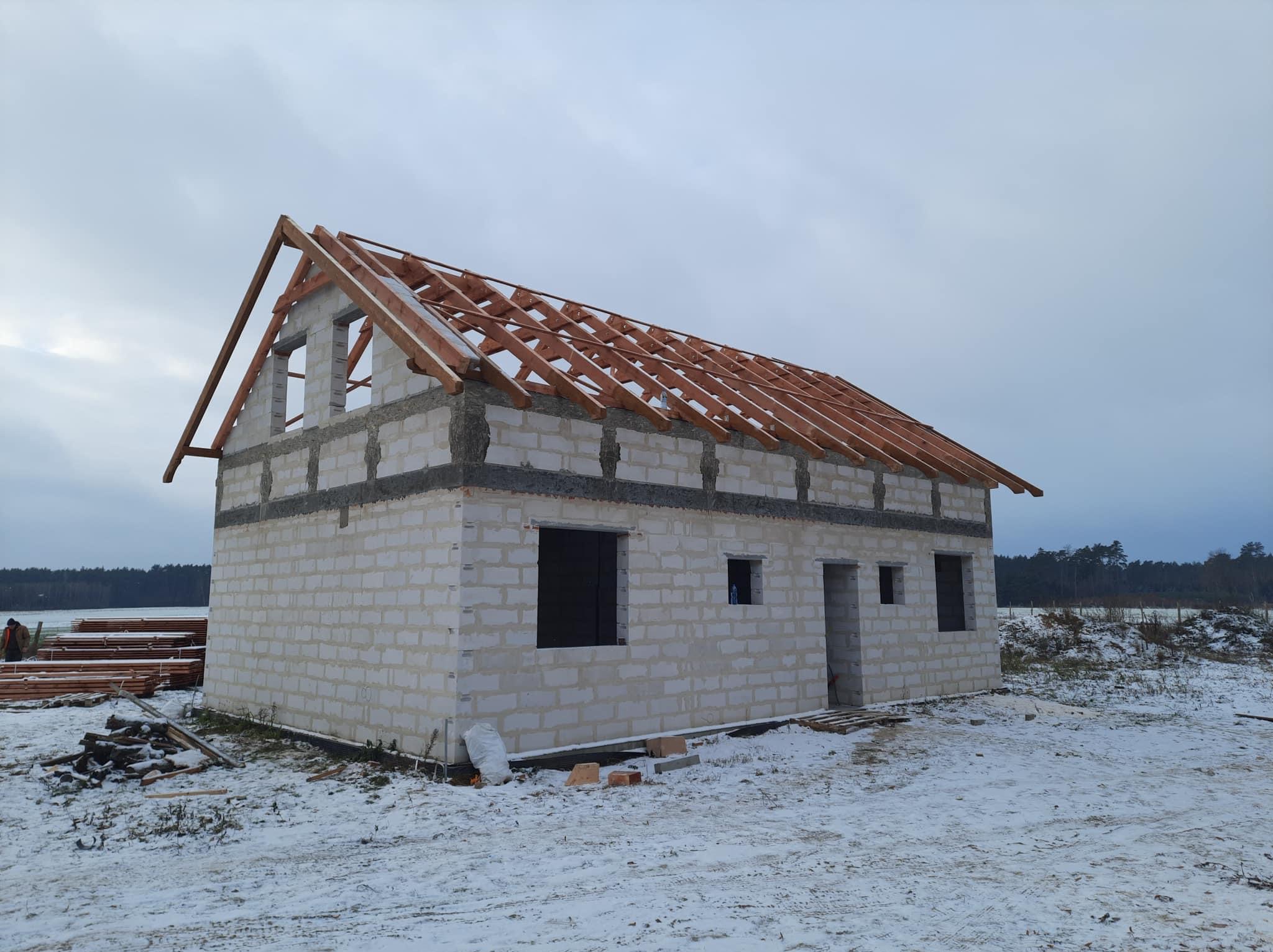 Niedokończony dom murowany z białych bloczków w zimowej scenerii, z drewnianą konstrukcją dachu bez pokrycia, widoczne stosy drewna i śnieg na ziemi.