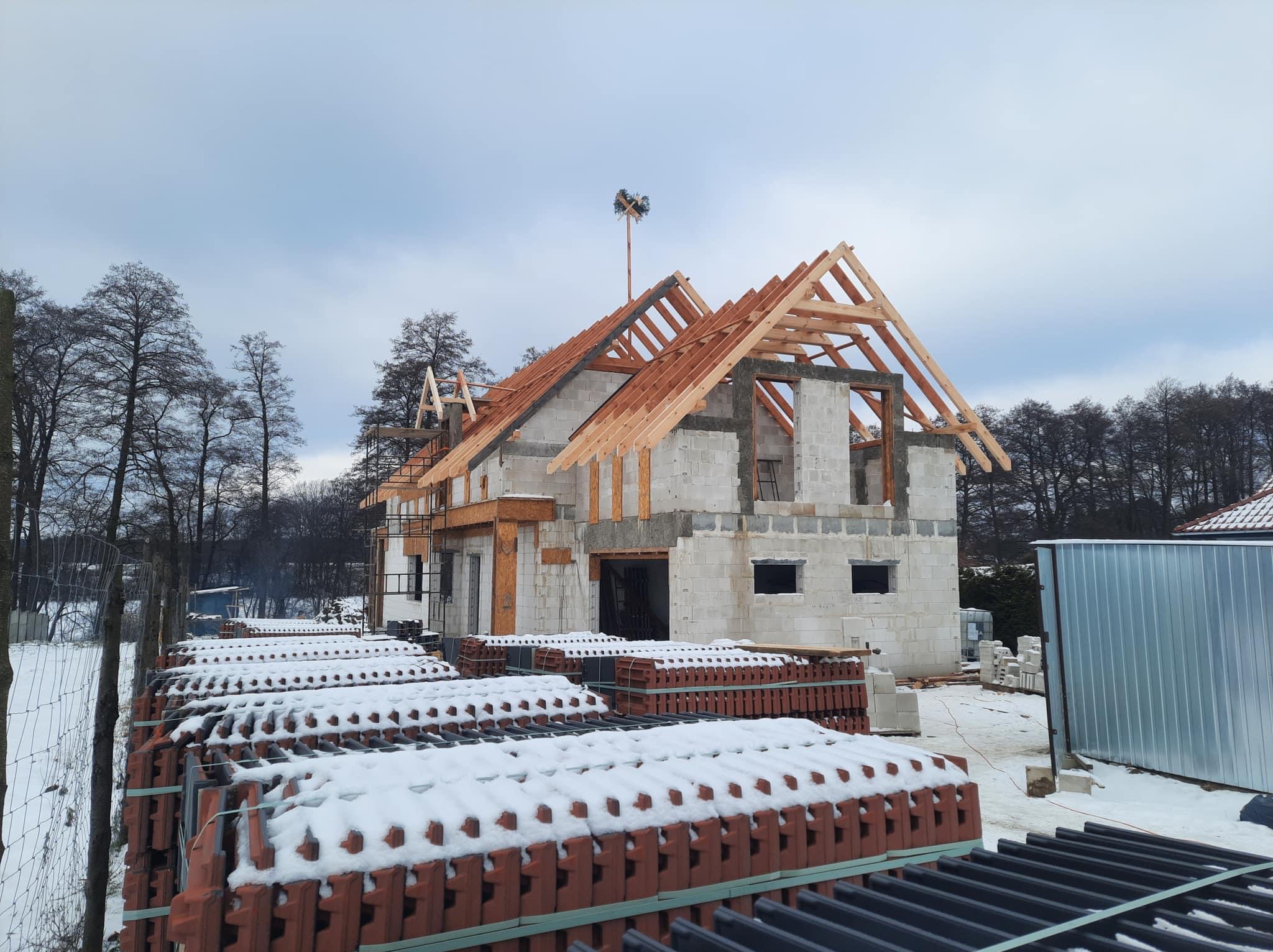 Surowy stan domu murowanego z drewnianą konstrukcją dachu w zimowej scenerii, palety czerwonej dachówki pokryte śniegiem na pierwszym planie.