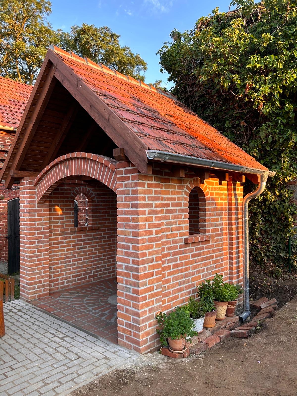 Murowana z cegły czerwonej altana z łukowymi oknami i dachem z dachówki ceramicznej, obok której stoją doniczki z roślinami na tle zieleni.