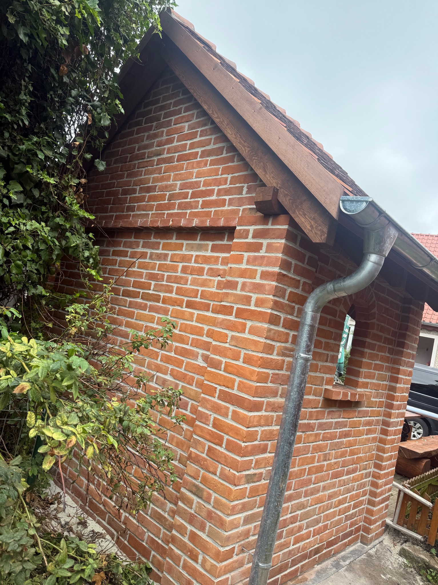 Fragment murowanej z czerwonej cegły dobudówki z drewnianą więźbą dachową i rynną, częściowo zasłoniętej zieloną roślinnością.