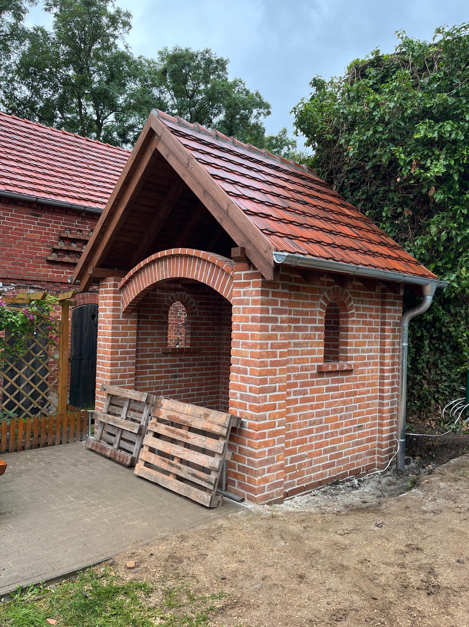 Murowana altana z czerwoną dachówką i drewnianą konstrukcją dachu. Widoczne palety obok konstrukcji. Tło stanowi zieleń drzew i krzewów.