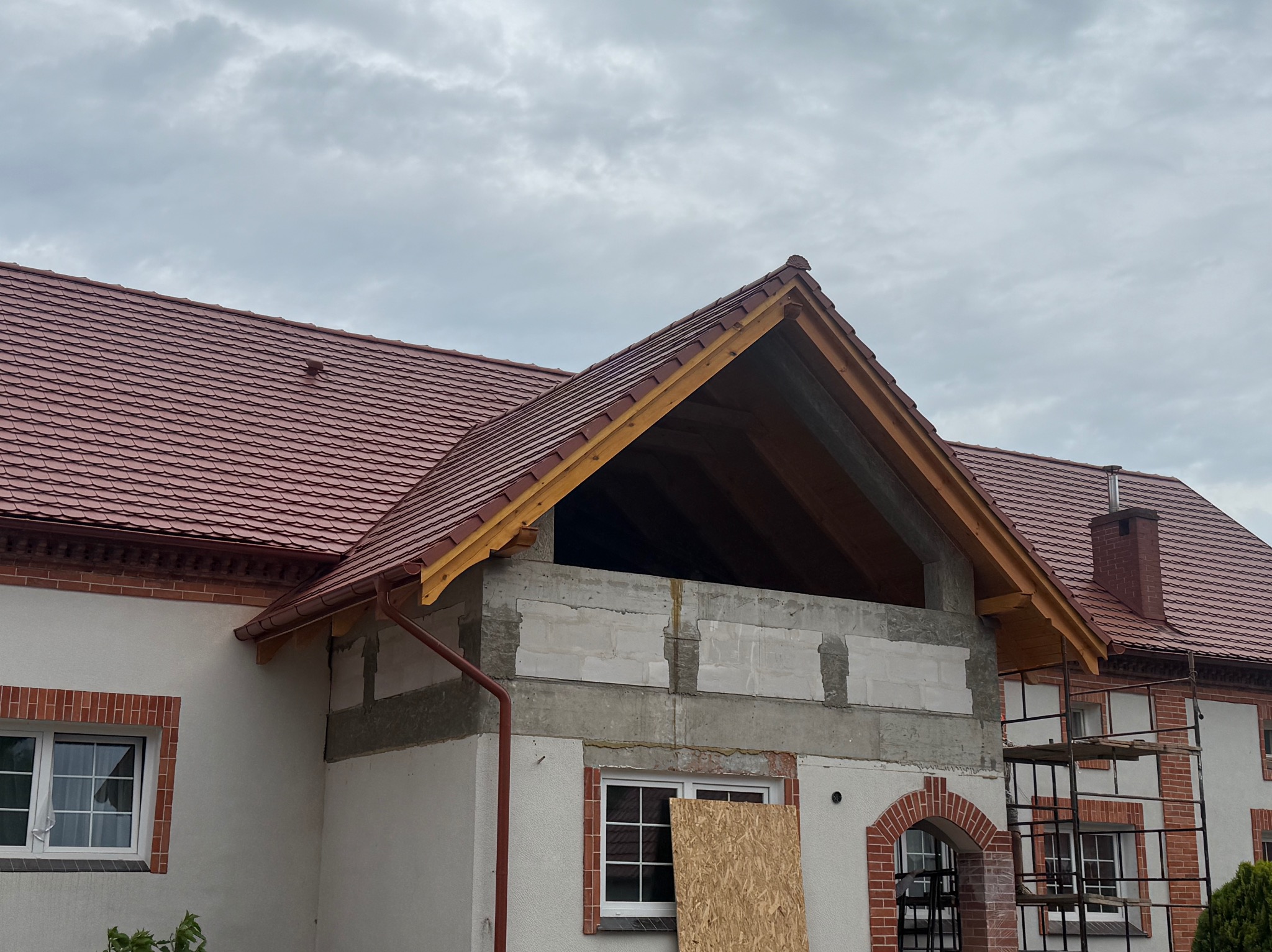 Rozbudowa domu z czerwoną dachówką, widoczna drewniana więźba dachowa i ściany z pustaków. Część okna zasłonięta płytą. Na drugim planie rusztowanie.