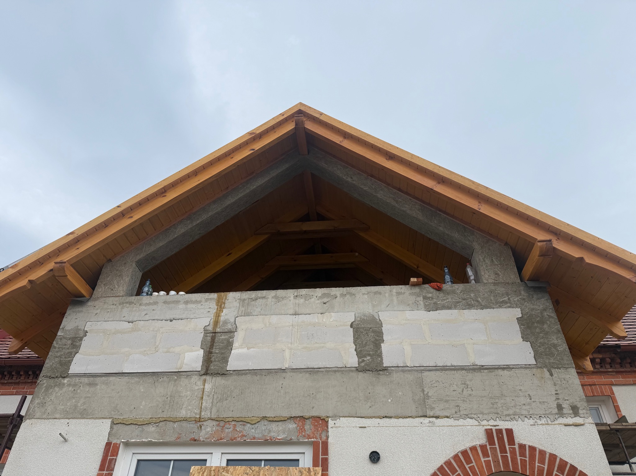 Wznoszenie ściany szczytowej z betonu komórkowego i drewnianej więźby dachowej. Widoczne niedokończone murowanie i elementy konstrukcyjne dachu.