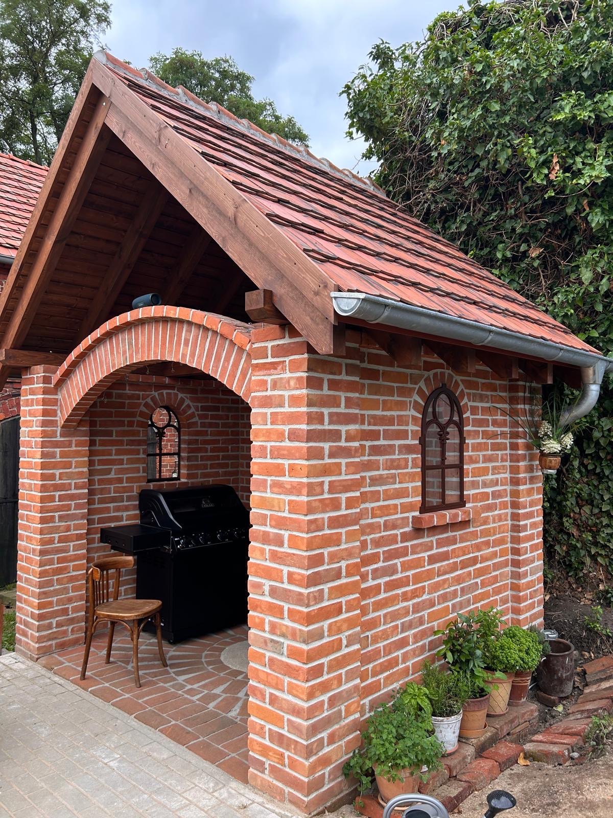 Murowana altana grillowa z czerwonej cegły, z drewnianą więźbą dachową i oknami w stylu retro. Na ceglanej podłodze stoi czarny grill i krzesło.