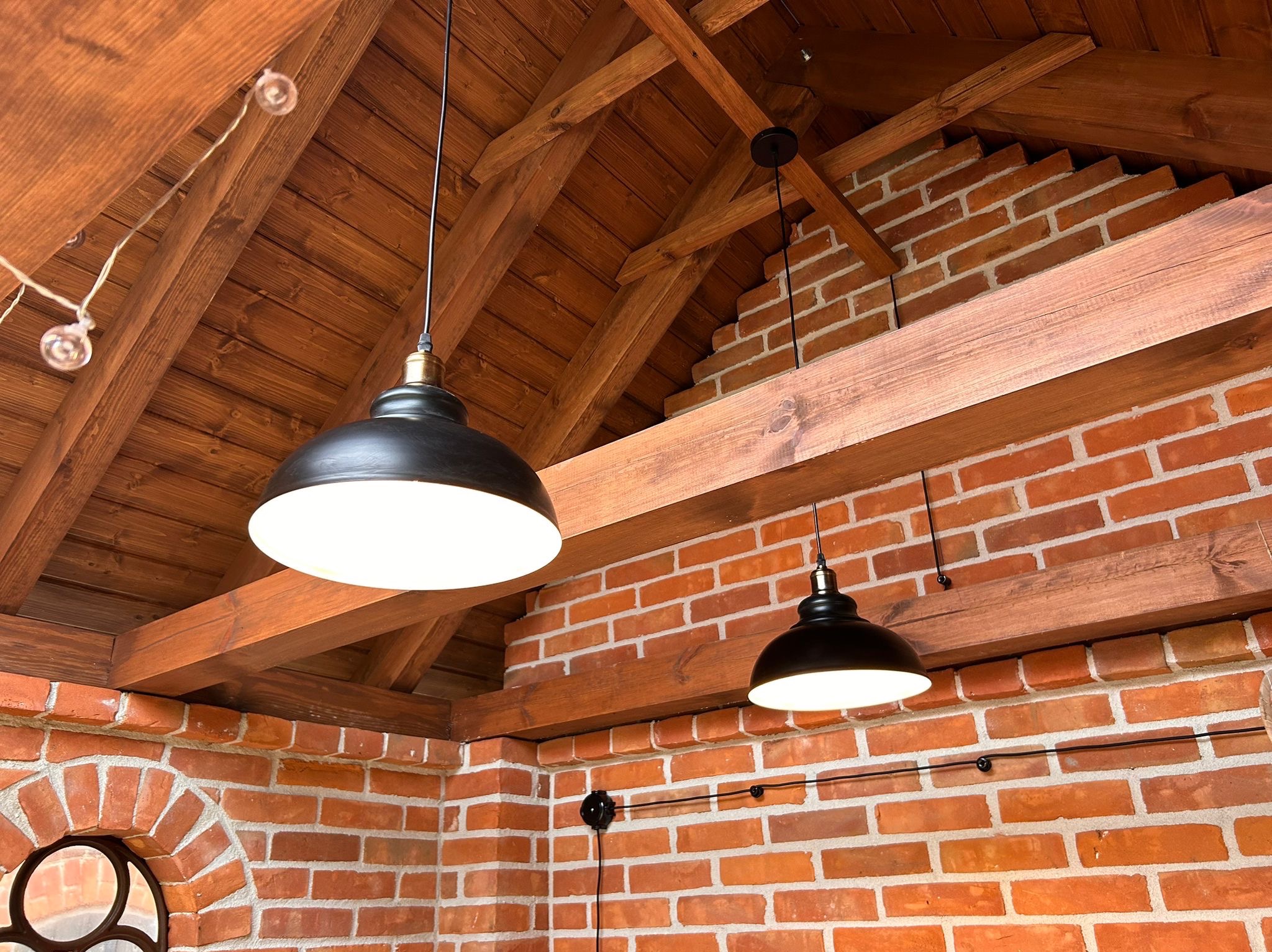 Ceglana ściana z drewnianą więźbą dachową, oświetlona dwoma czarnymi lampami wiszącymi. Widoczne detale instalacji elektrycznej i dekoracyjne oświetlenie.