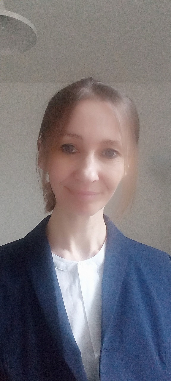 Portret kobiety w białej koszuli i granatowej marynarce, uśmiechająca się, z widoczną lampą sufitową w tle.