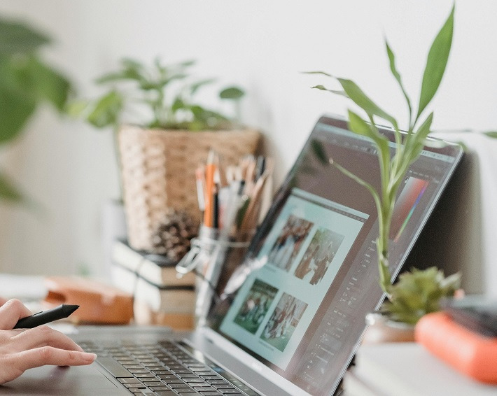 Dłoń z piórkiem graficznym retuszuje zdjęcia ślubne w programie graficznym na laptopie, w tle rośliny doniczkowe i przybory do pisania.