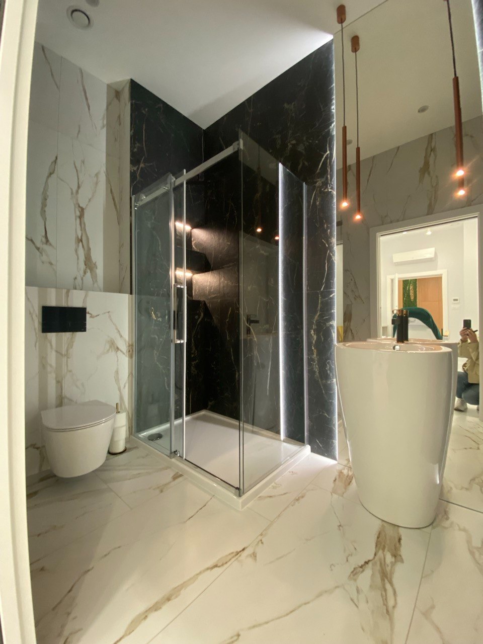 Nowoczesna łazienka z kabiną prysznicową typu walk-in, umywalką wolnostojącą i podwieszaną toaletą, wykończona płytkami imitującymi marmur w kolorze białym i czarnym ze złotymi żyłami.