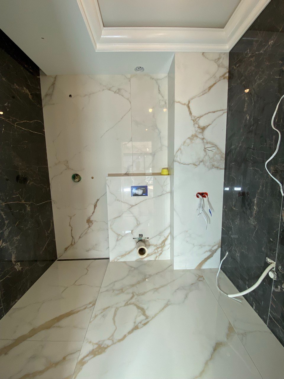 Wnętrze łazienki w trakcie prac wykończeniowych, widoczne białe płytki imitujące marmur z złotymi żyłami na ścianach i podłodze, czarne płytki po bokach, przygotowane przyłącze do toalety...