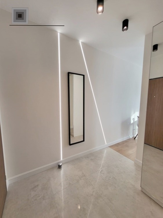 Nowoczesny przedpokój z ukrytym oświetleniem LED wzdłuż ściany, lustro w czarnej ramie, szare płytki podłogowe z połyskiem i minimalistyczne czarne reflektory sufitowe.