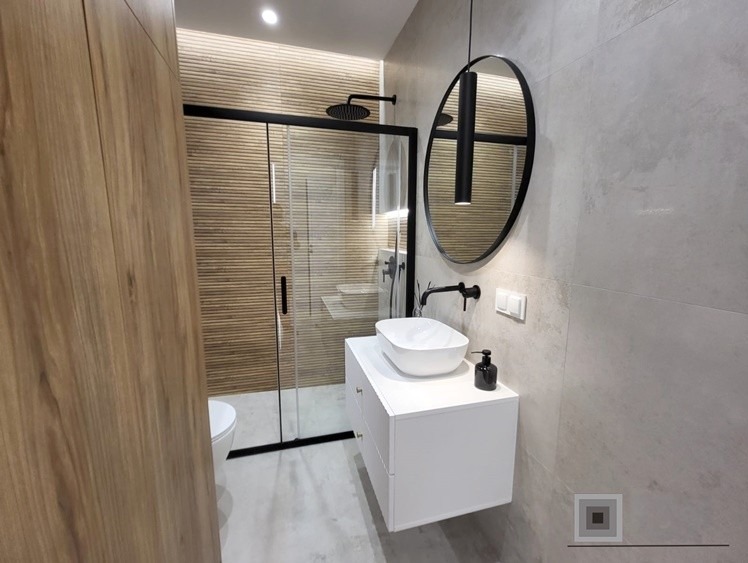 Nowoczesna łazienka z kabiną prysznicową typu walk-in, umywalką nablatową na białej szafce, okrągłym lustrem z oświetleniem oraz płytkami imitującymi drewno na ścianie w kabinie.