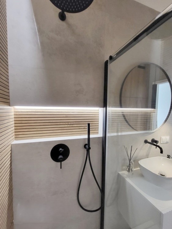 Nowoczesna łazienka z kabiną prysznicową typu walk-in, czarną armaturą, betonową wylewką na ścianach, podświetlanymi drewnianymi panelami i okrągłym lustrem.