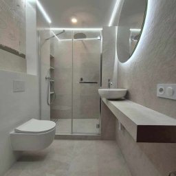 Magkambud - Nowoczesna łazienka w odcieniach szarości z podwieszaną toaletą, kabiną prysznicową ze szklanymi drzwiami, umywalką nablatową na betonowym blacie i okrągłym lustrem z podświetleniem LED.