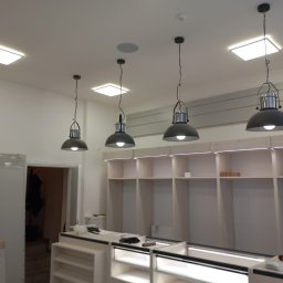 Magkambud - Montaż oświetlenia w nowym biurze: cztery czarne lampy wiszące nad jasnymi regałami z podświetleniem LED, widoczne również kwadratowe lampy sufitowe.