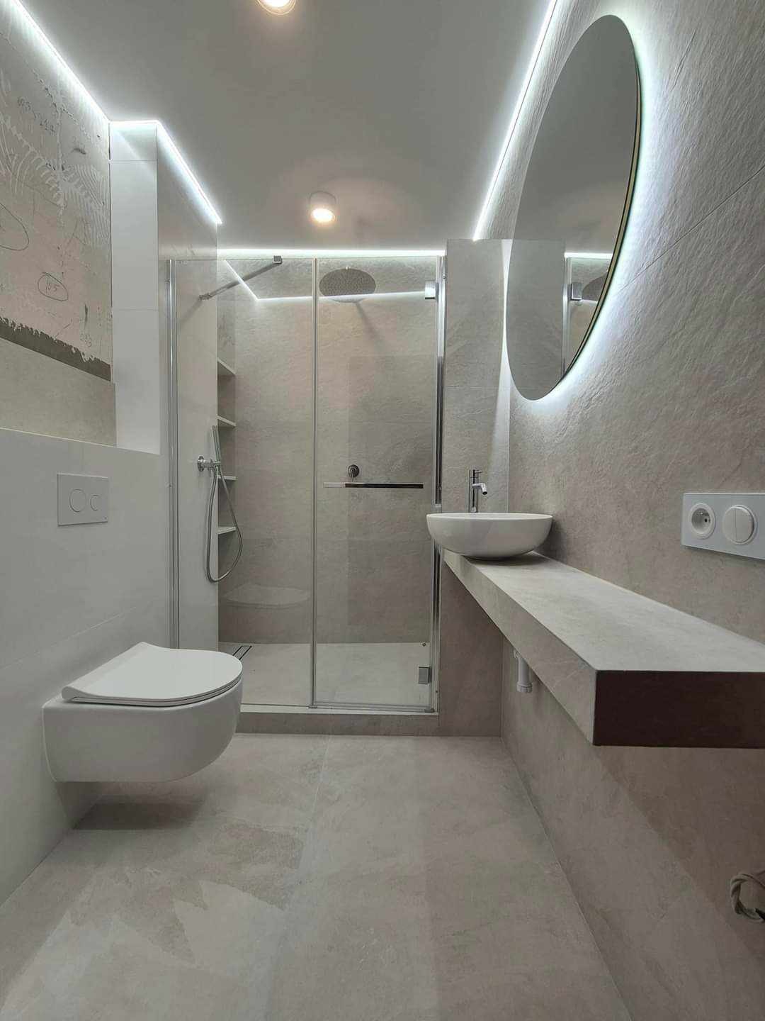 Nowoczesna łazienka w odcieniach szarości z podwieszaną toaletą, kabiną prysznicową ze szklanymi drzwiami, umywalką nablatową na betonowym blacie i okrągłym lustrem z podświetleniem LED.