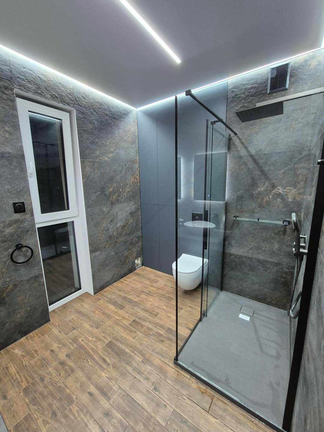Nowoczesna łazienka z szarymi płytkami imitującymi kamień, drewnopodobną podłogą, kabiną prysznicową typu walk-in z czarnymi profilami i oświetleniem LED w suficie.