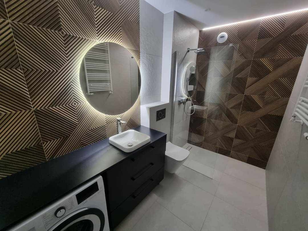 Nowoczesna łazienka z okrągłym lustrem podświetlanym LED, drewnianą mozaiką na ścianach, czarną szafką pod umywalką i pralką, kabiną prysznicową za szkłem.