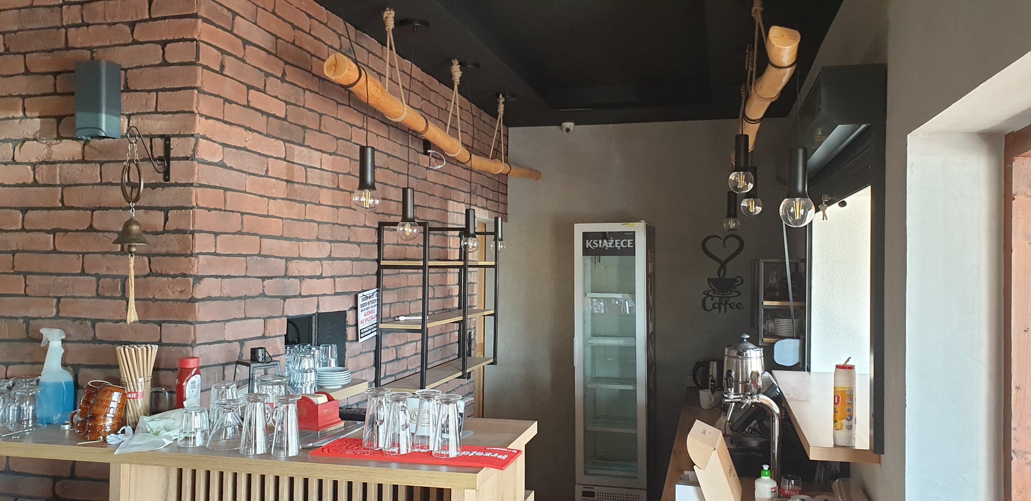 Wnętrze kawiarni z ceglaną ścianą, drewnianymi lampami, półkami, szklankami na barze i lodówką z napojami
