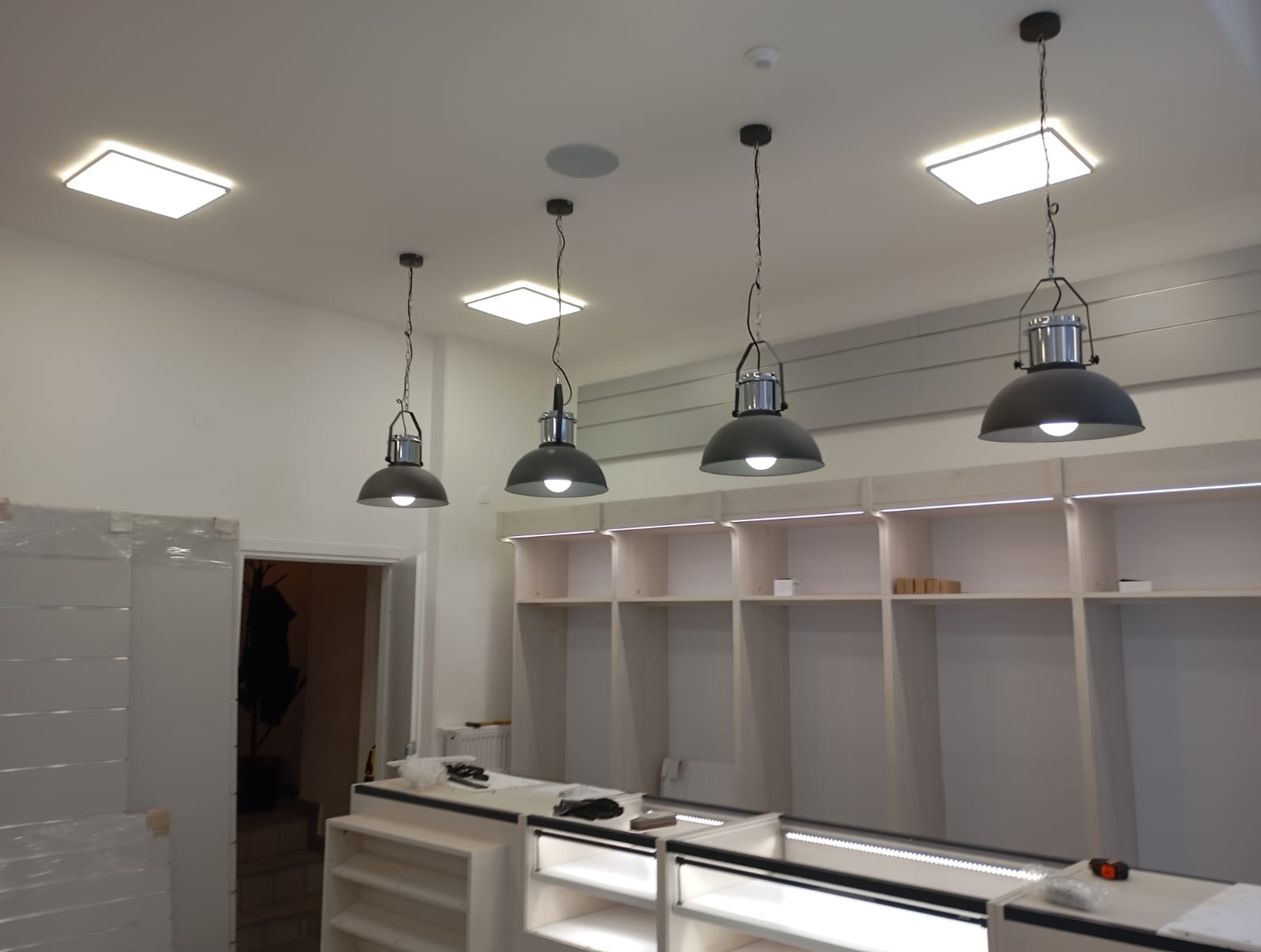 Montaż oświetlenia w nowym biurze: cztery czarne lampy wiszące nad jasnymi regałami z podświetleniem LED, widoczne również kwadratowe lampy sufitowe.