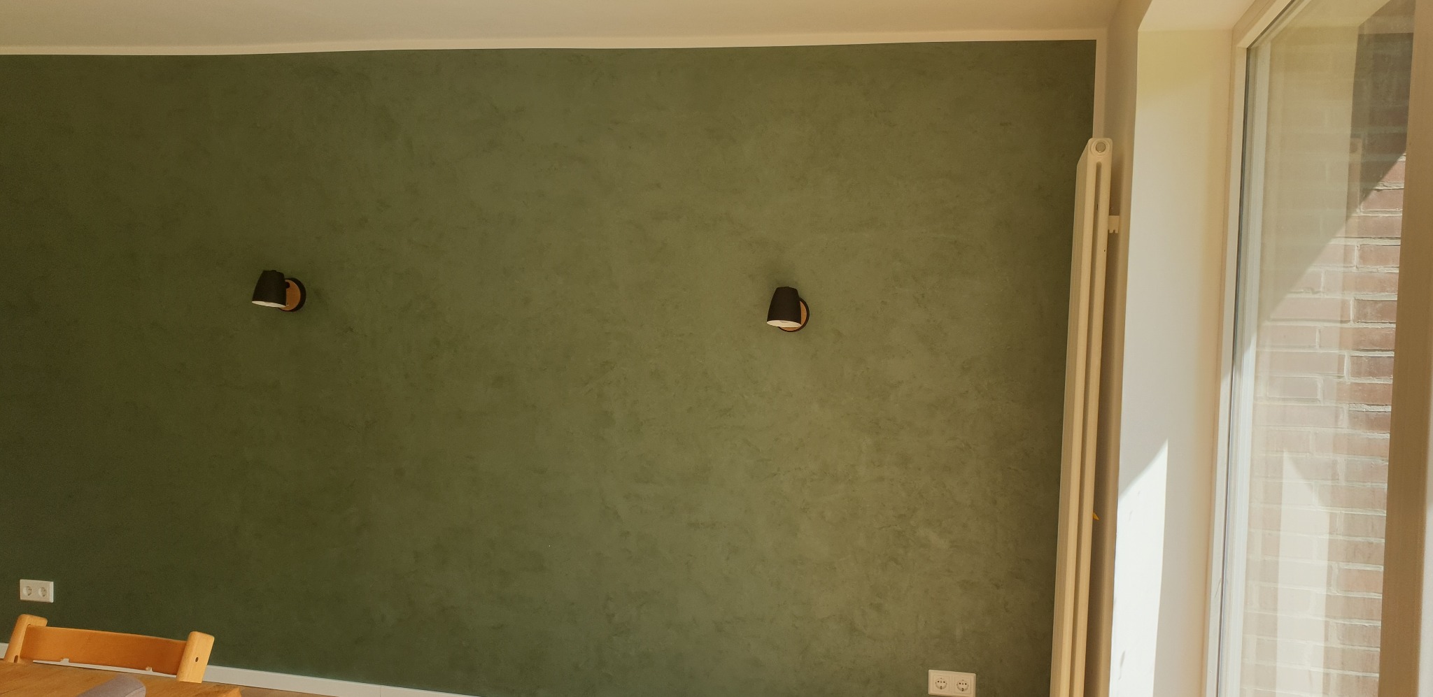 Ściana w odcieniach zieleni z dekoracyjną fakturą, oświetlona dwoma kinkietami i światłem dziennym wpadającym przez okno, widoczny fragment grzejnika i gniazdka elektryczne.