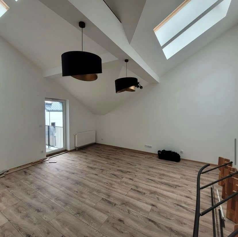 Pokój na poddaszu z nową podłogą z paneli w odcieniach brązu, świeżo pomalowane ściany na biało, czarne, okrągłe lampy wiszące i okno dachowe.