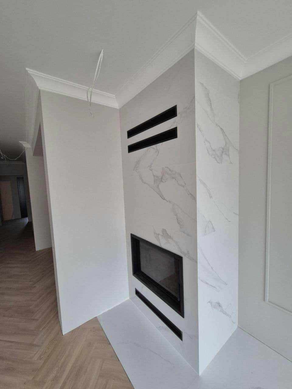 Obudowa kominka z białych płytek imitujących marmur, z czarnymi akcentami, w nowym wnętrzu z parkietem w jodełkę.