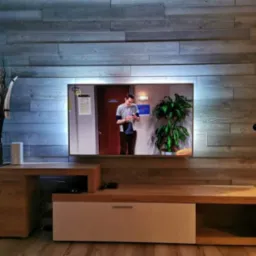 Ściana telewizyjna wyłożona panelami imitującymi drewno, podświetlona od tyłu, z szafką RTV w kolorze drewna i białym frontem, widoczny telewizor z włączonym serialem.