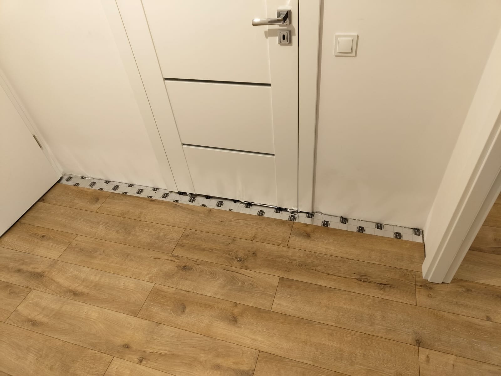 Jasne panele podłogowe imitujące drewno, ułożone wzdłuż białej ściany i ościeżnicy drzwiowej, widoczne paski folii ochronnej na styku podłogi ze ścianą.