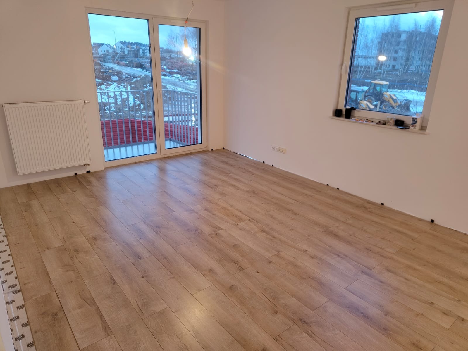 Układanie paneli podłogowych w jasnym, nowym mieszkaniu z widokiem na plac budowy za oknem.