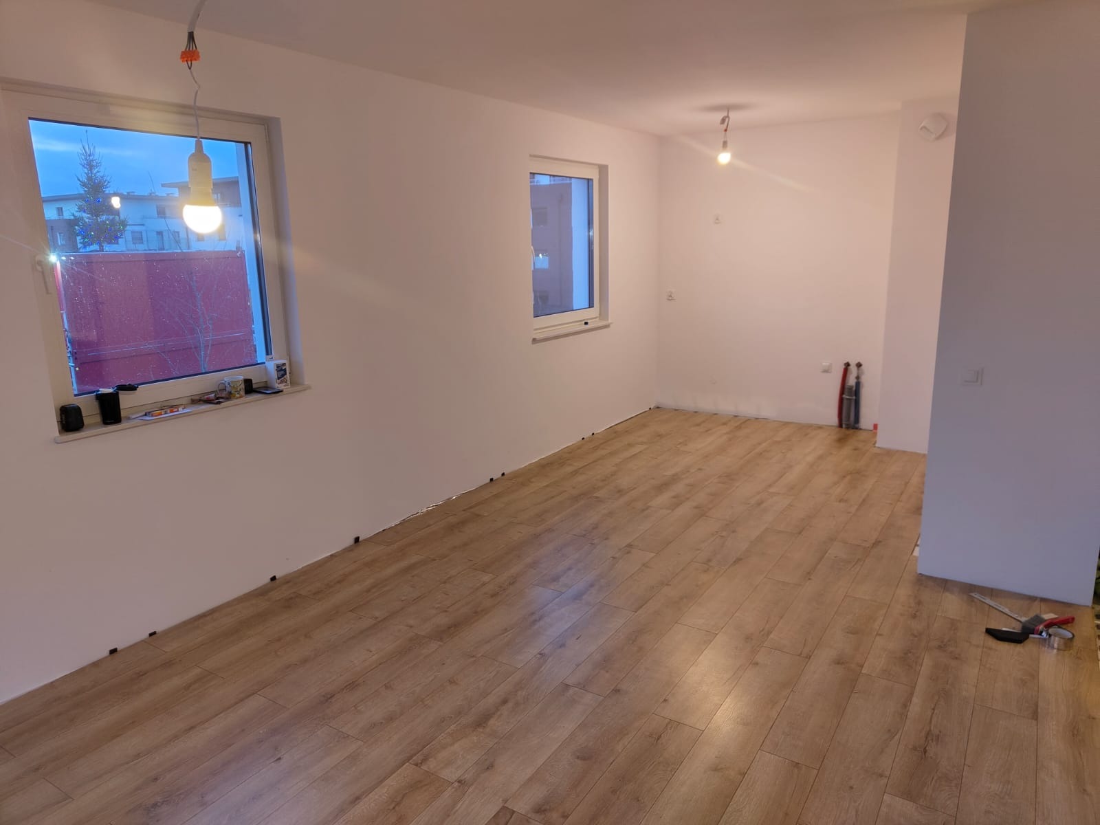 Pokój z częściowo ułożoną podłogą z paneli laminowanych w odcieniach drewna, widoczne narzędzia i listwy przypodłogowe, okno z widokiem na zewnątrz.