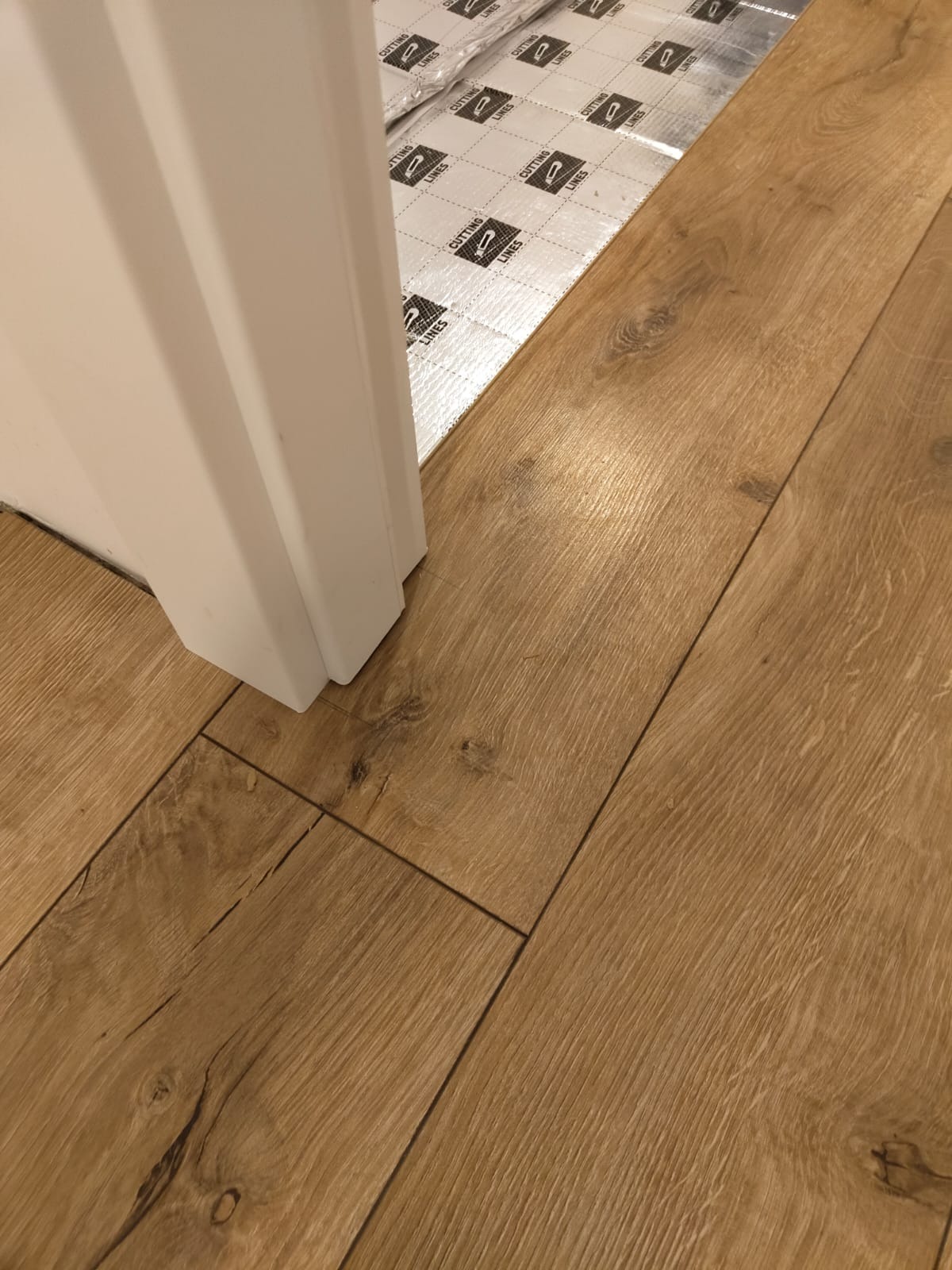 Fragment podłogi z paneli w odcieniach dębu, widoczne podłoże izolacyjne z nadrukowanymi liniami cięcia, tuż przy białej ościeżnicy drzwiowej.