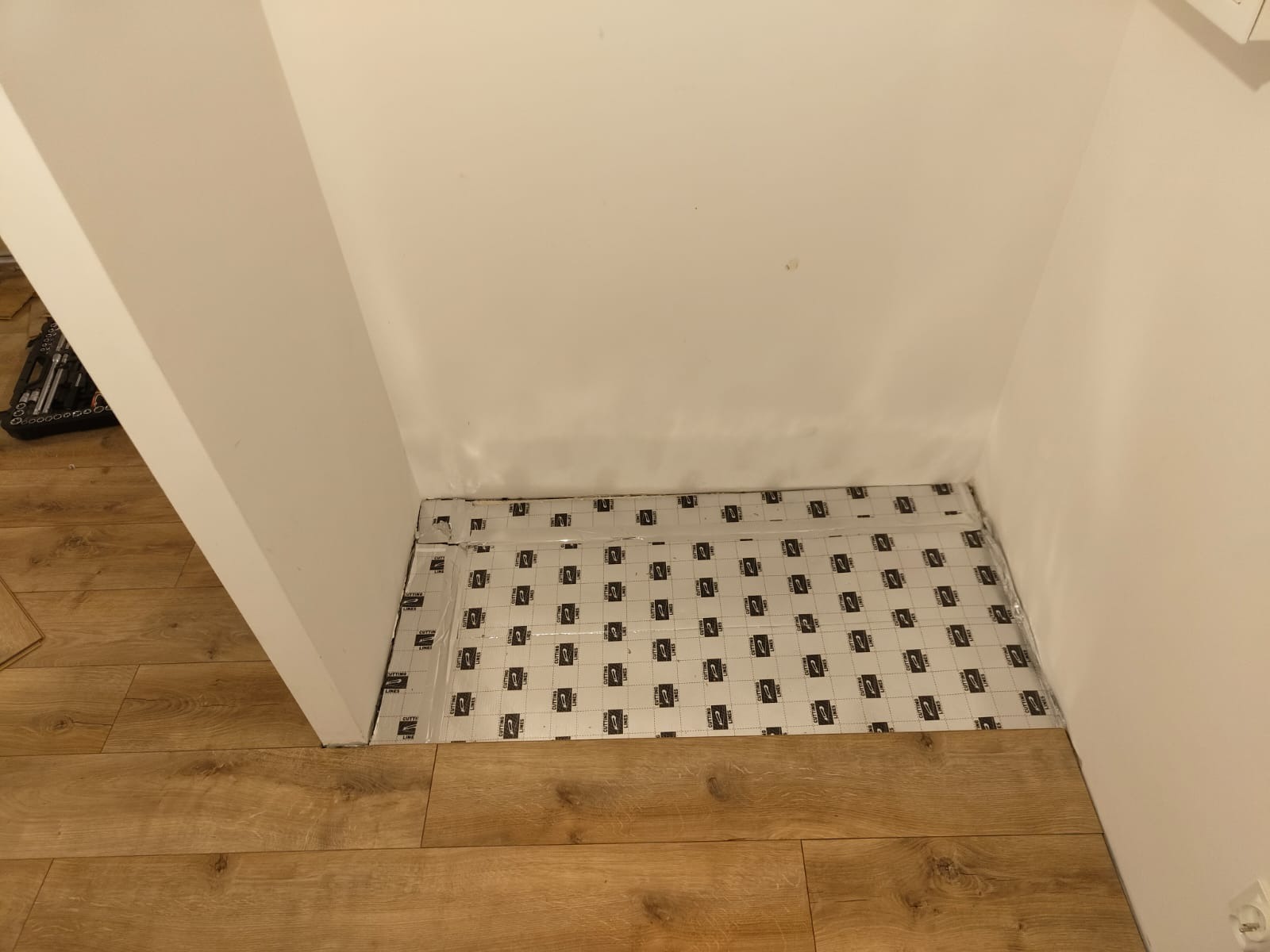 Przygotowanie podłoża pod panele: rozłożona folia izolacyjna z nadrukiem w małym wnęku pomieszczenia, widoczna część drewnopodobnej podłogi i narzędzia.