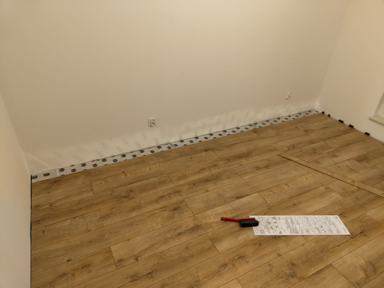Fragment pokoju z częściowo ułożoną podłogą z paneli w odcieniach brązu, widoczne podkładki pod panele przy ścianach, instrukcja montażu i szczotka do zamiatania na ułożonej powierzchni.