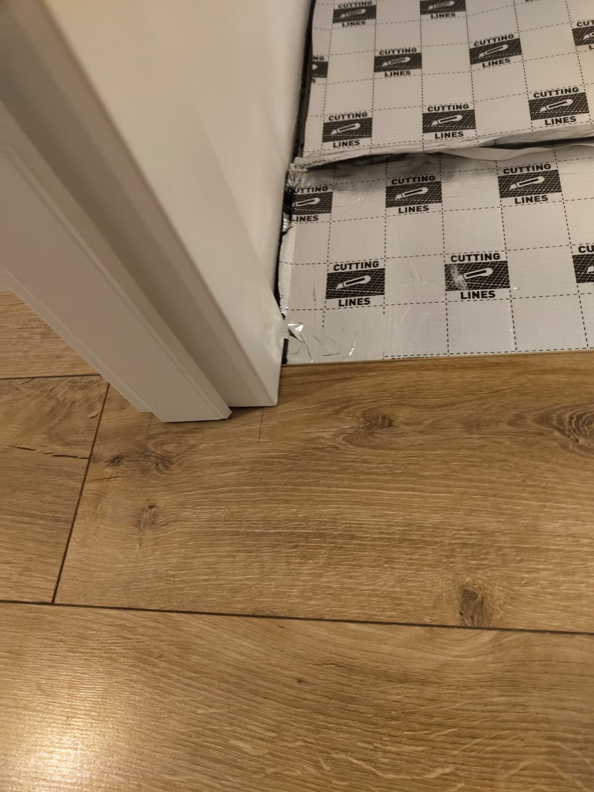 Przycięta mata ochronna z nadrukiem 'CUTTING LINES' pod białą ościeżnicą, na drewnianej podłodze z paneli.