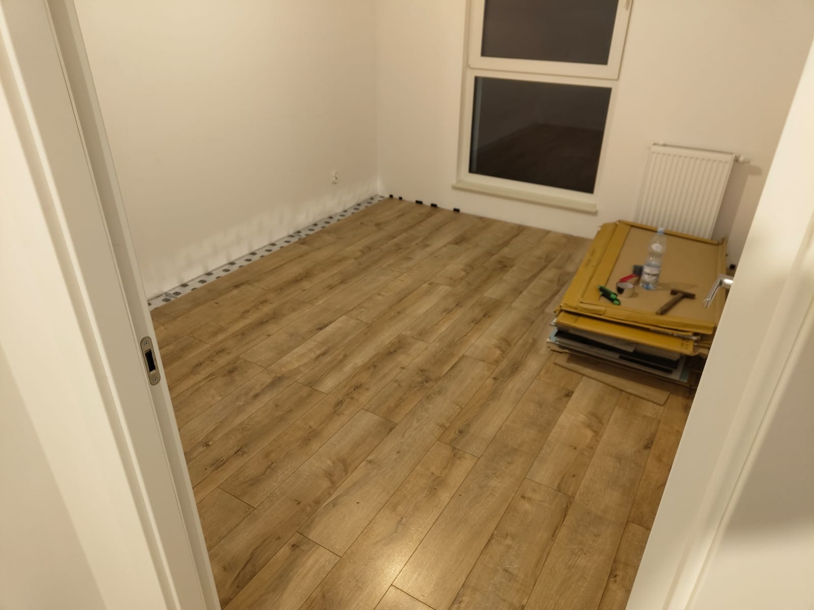 Układanie paneli podłogowych w pokoju, widoczna podkładka izolacyjna przy ścianie, ułożona część paneli, narzędzia i panele czekające na montaż.