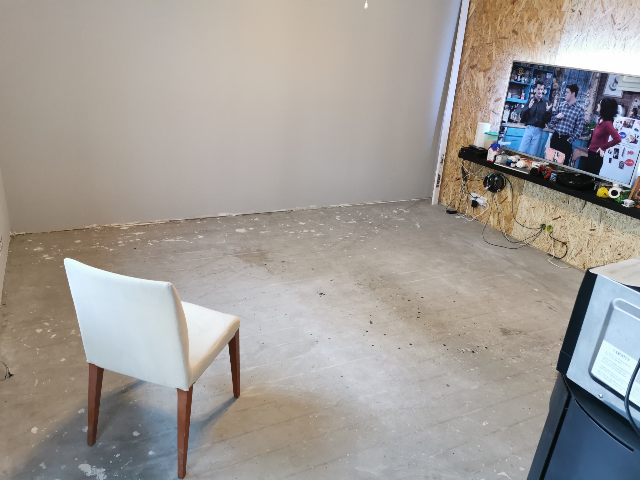 Przygotowanie do układania podłogi: gołe podłoże z widocznymi śladami po kleju i farbie, białe krzesło na środku i telewizor z serialem 'Przyjaciele' w tle.