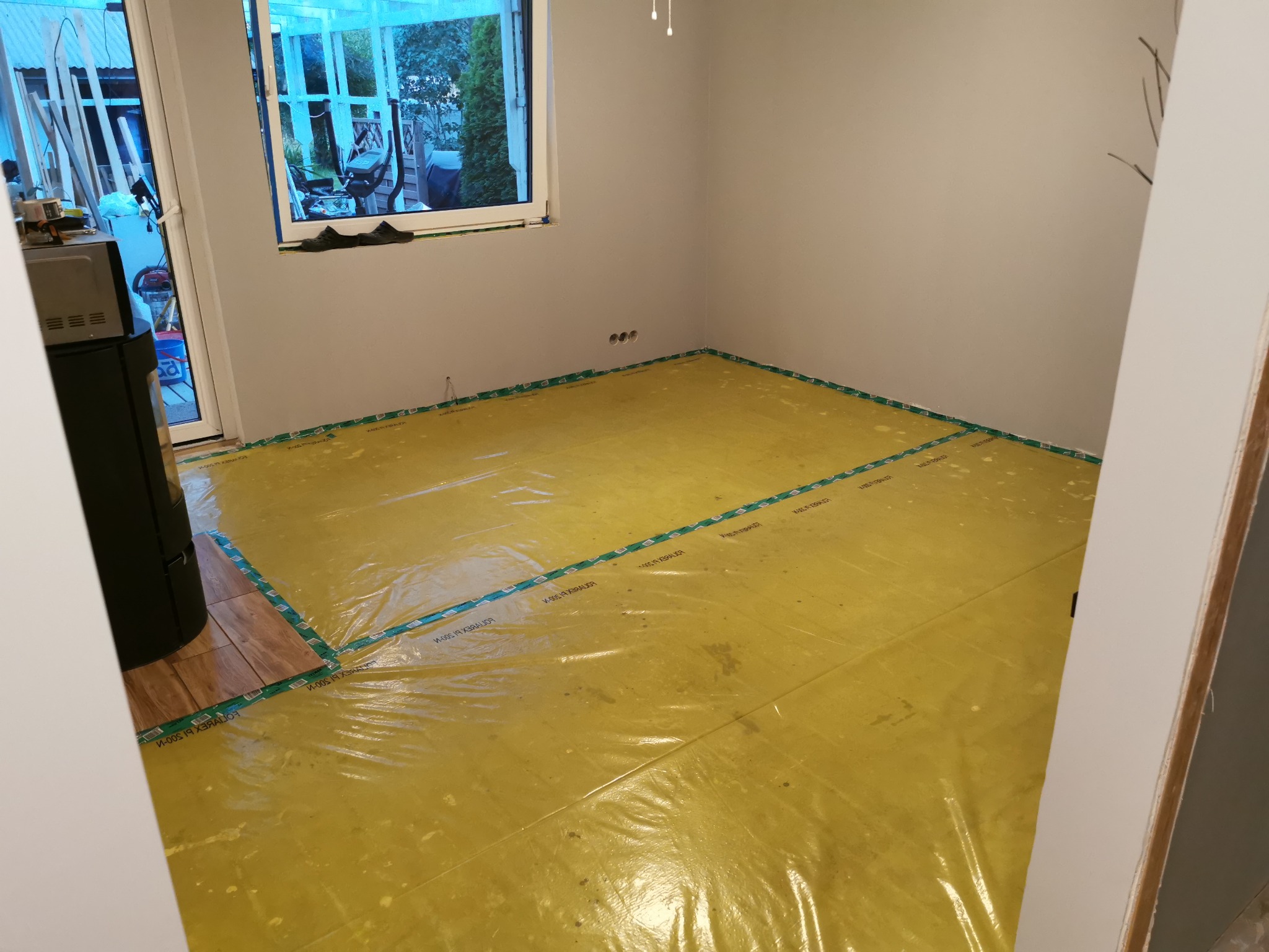 Przygotowanie podłoża pod montaż podłogi: rozłożona folia ochronna z zieloną taśmą brzegową w pustym pomieszczeniu z szarymi ścianami i widocznymi gniazdkami elektrycznymi, częściowo ułożone panele...