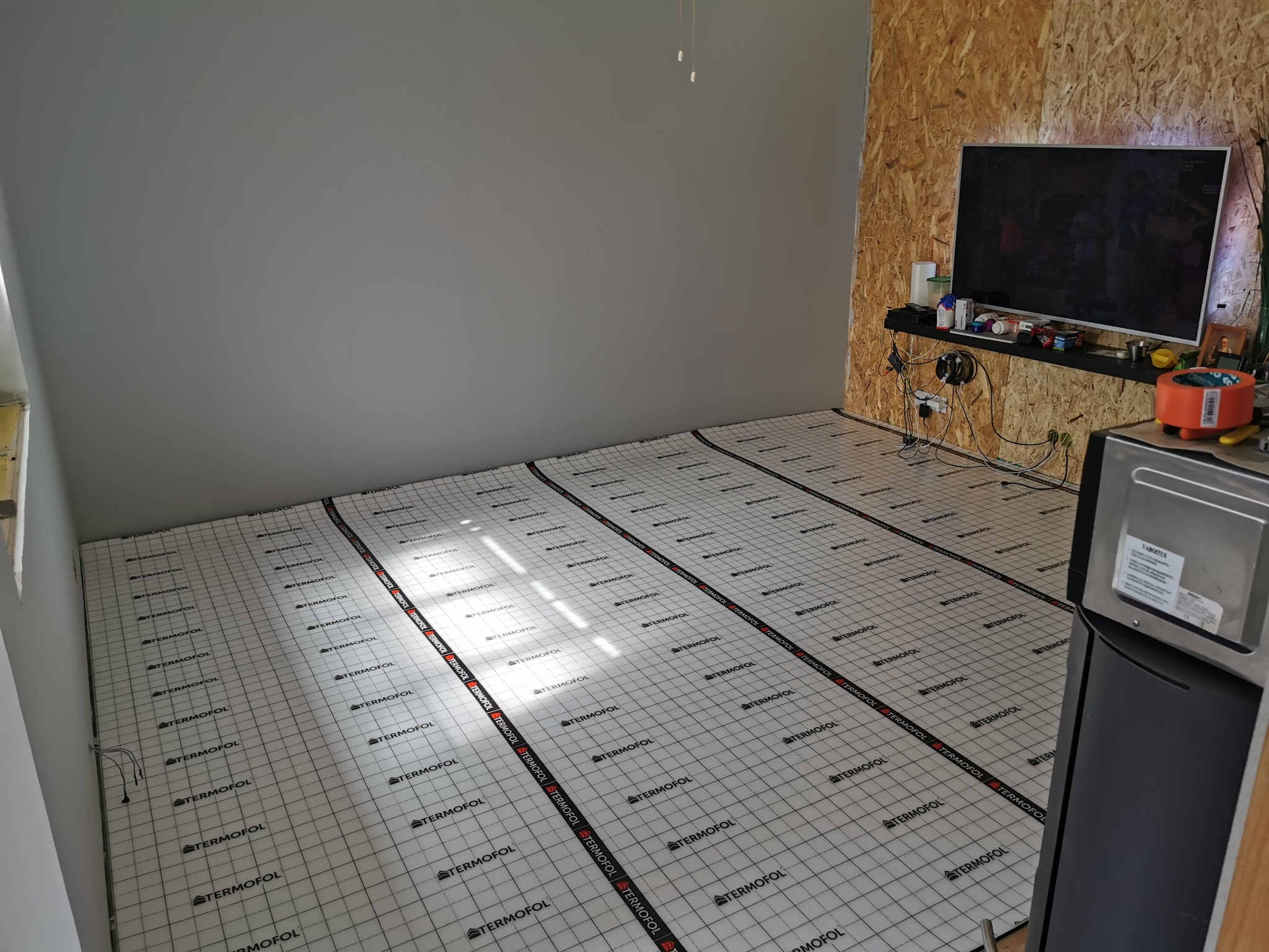 Przygotowanie podłoża pod panele podłogowe: rozłożona mata izolacyjna Termofol z nadrukowaną siatką na wylewce w pokoju z szarą ścianą i ścianą wykończoną płytą OSB.