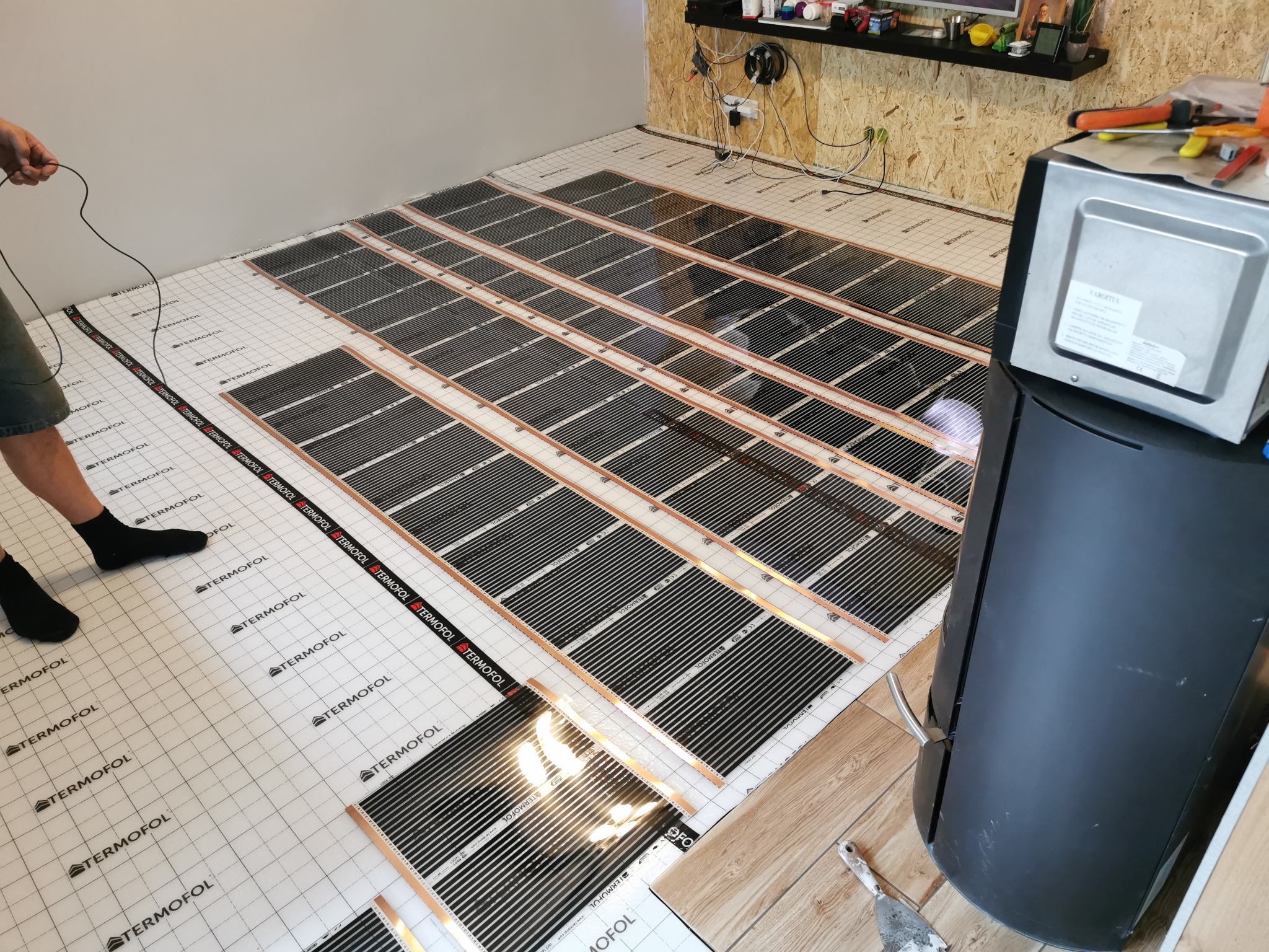 Montaż folii grzewczej pod panele podłogowe; widoczne czarne maty grzewcze firmy Termofol rozłożone na białej siatce z nadrukiem, fragment nogi instalatora w czarnej skarpetce, oraz piec...