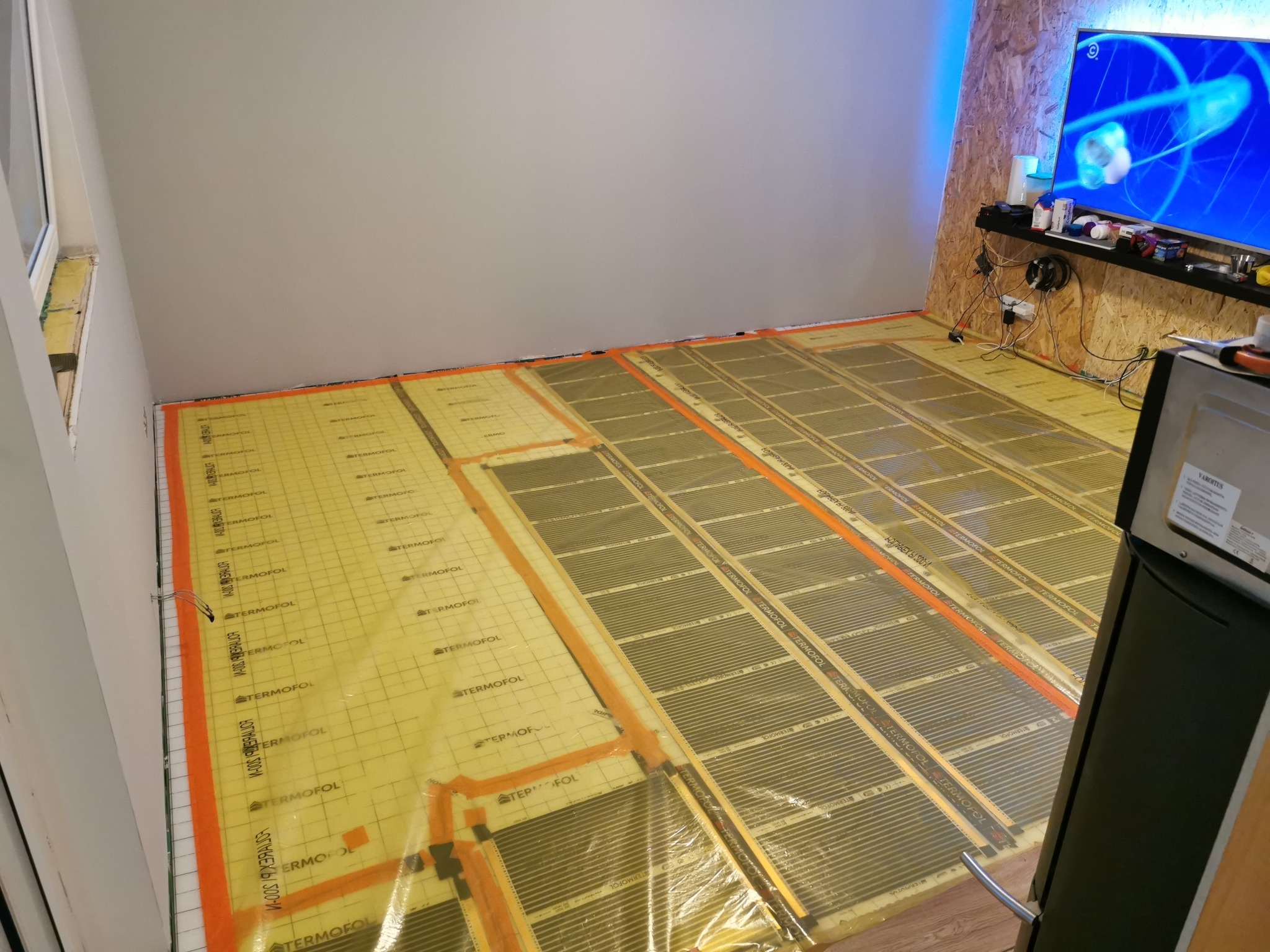 Układanie folii grzewczej Termofol na podłodze, widoczne paski grzewcze z pomarańczowymi krawędziami, przygotowanie do montażu paneli podłogowych w pokoju z telewizorem.