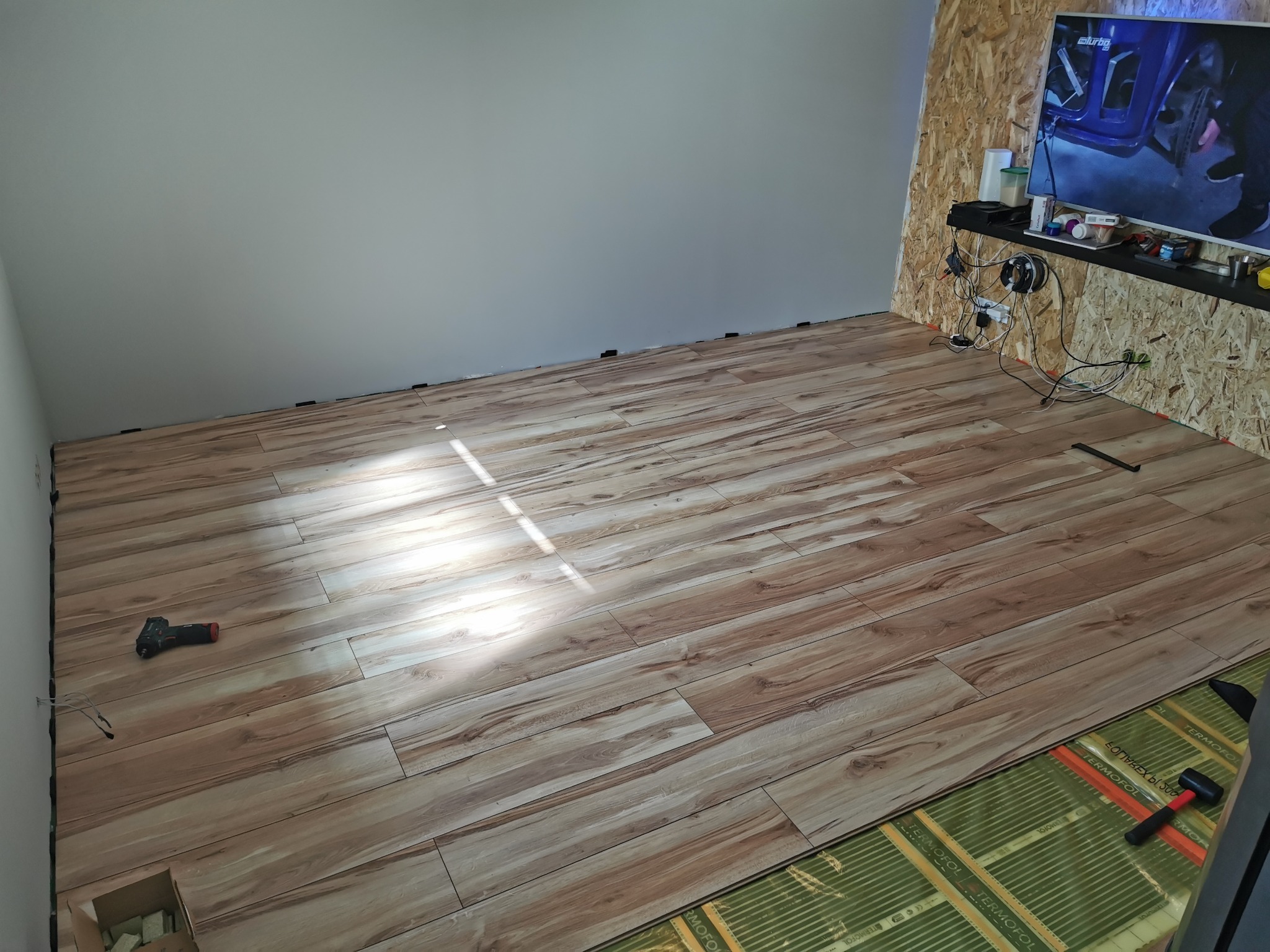 Fragment pokoju z panelami podłogowymi o wzorze drewna, częściowo ułożonymi na zielonej macie izolacyjnej, widoczne narzędzia: wkrętarka i młotek gumowy.