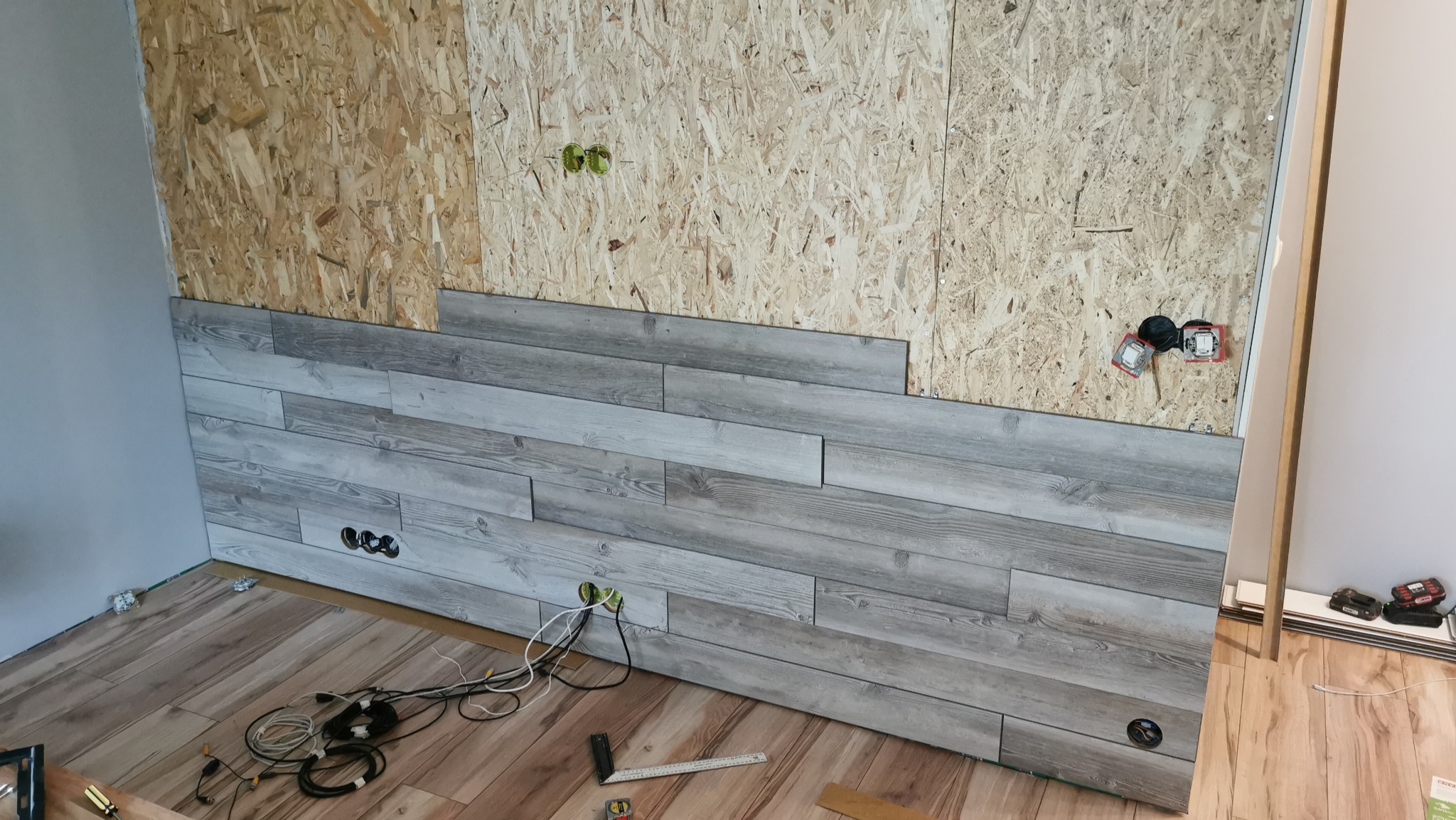 Fragment ściany z częściowo ułożonymi panelami ściennymi o wzorze imitującym drewno, widoczne gniazdka elektryczne, narzędzia pomiarowe i płyta OSB w górnej części ściany.
