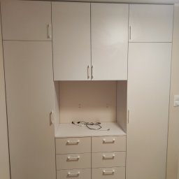 Kamil Wąsik - Biała zabudowa meblowa z szafkami górnymi i dolnymi z szufladami, z otwartą przestrzenią na blacie z widocznymi kablami, na podłodze parkiet ułożony w jodełkę.