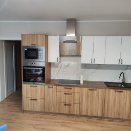 Kamil Wąsik - Nowoczesna kuchnia z frontami w kolorze drewna i białymi szafkami górnymi, czarne uchwyty, marmurowy backsplash, sprzęt AGD zabudowany w wysokiej szafce, płyta indukcyjna i czarny zlew.