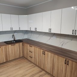 Kamil Wąsik - Nowoczesna kuchnia z frontami w kolorze jasnego drewna i białymi szafkami górnymi z czarnymi uchwytami; blat w ciemnym odcieniu brązu, ściana nad blatem wykończona płytkami imitującymi marmur.